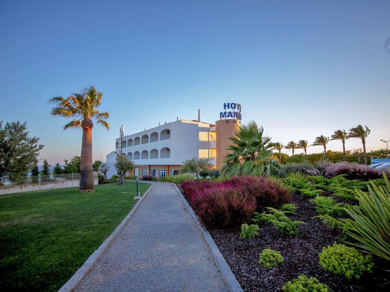 Ein Hotelgebäude mit weißen Balkonen, umgeben von gepflegtem Garten mit Palmen und Pflanzen.