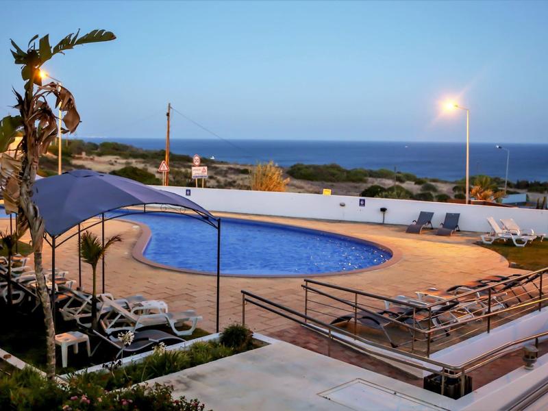 Schwimmbad mit blauer Wasserfläche, Liegestühlen, Palmen und Blick auf Meer bei Abenddämmerung.