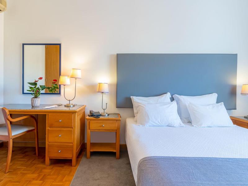 Helles Schlafzimmer mit blaugrauem Kopfteil, weißer Bettwäsche, Holzmöbeln und warmem Licht.