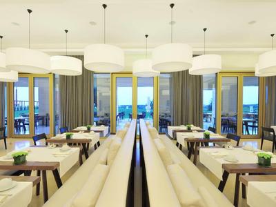 Modernes Restaurant mit weißen Lampen, Holztischen, Stühlen und Panoramafenstern mit Meerblick.