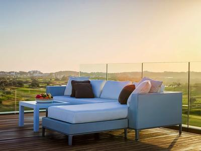 Blaues Sofa mit bunten Kissen auf Holzterrasse und Glasgeländer bei Sonnenuntergang.