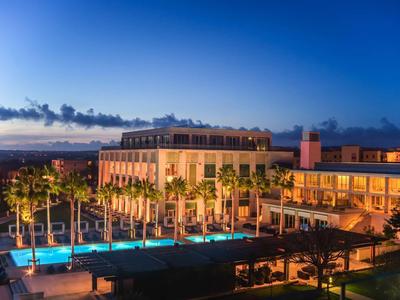 Großes Hotelgebäude mit beleuchtetem Pool und Palmen bei Sonnenuntergang unter blauem Himmel.