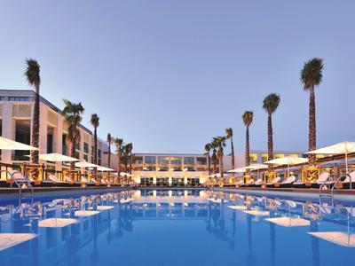 Ein großer blauer Pool mit Palmen und sonnengelben Sonnenschirmen zwischen weißen Gebäuden.