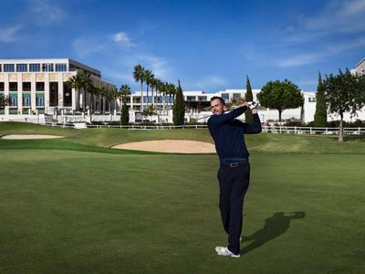 Mann spielt Golf auf gepflegtem Rasen vor Hotelgebäuden unter klarem Himmel.