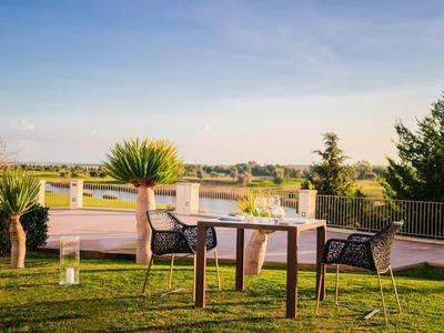 Tisch mit zwei Stühlen auf grüner Wiese, umgeben von Pflanzen und weitem Ausblick unter blauem Himmel.