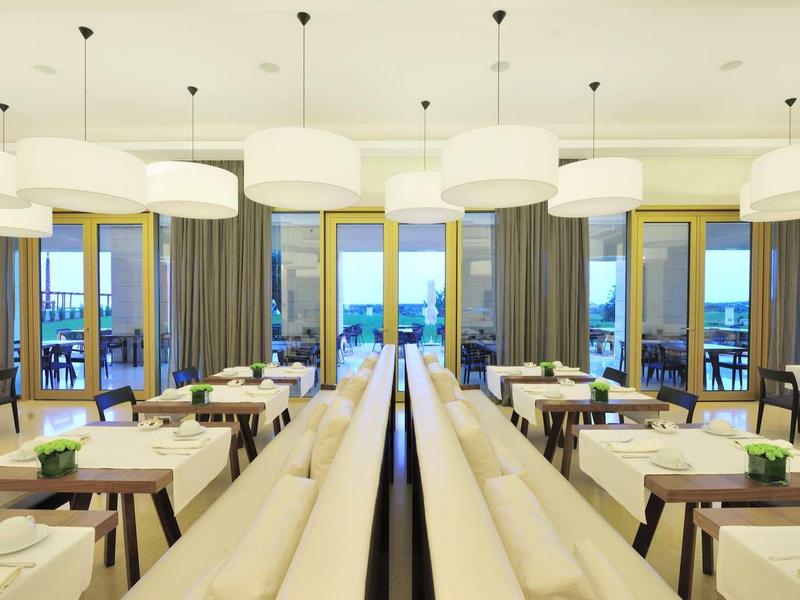 Modernes Restaurant mit weißen Lampen, Holztischen, Stühlen und Panoramafenstern mit Meerblick.