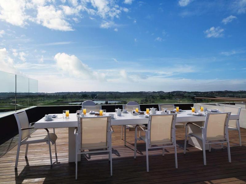Großer Esstisch mit Stühlen auf Holzterrasse, blauer Himmel und weiter Blick auf Felder.