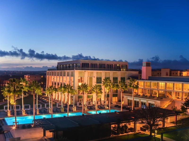 Großes Hotelgebäude mit beleuchtetem Pool und Palmen bei Sonnenuntergang unter blauem Himmel.
