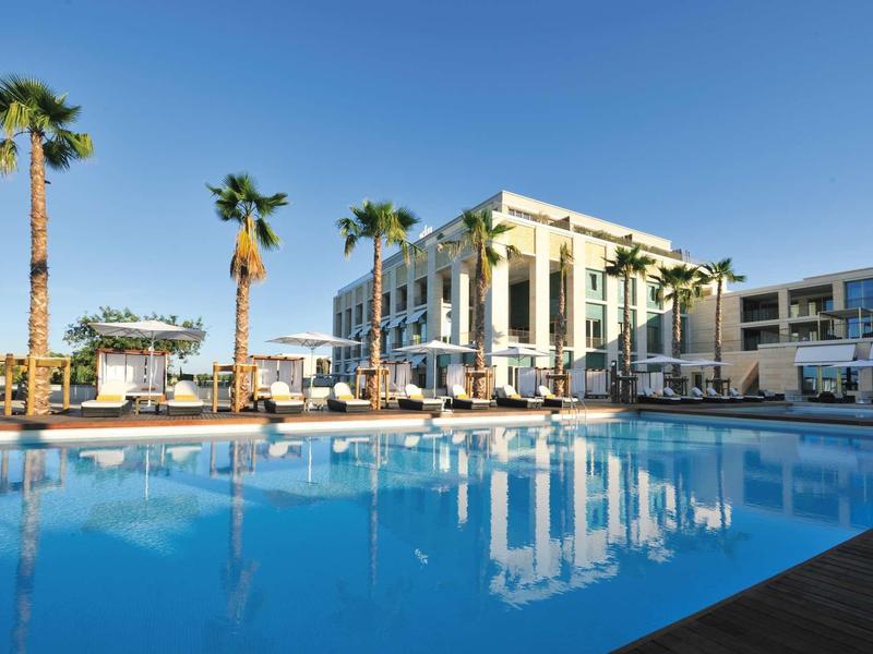 Großer blauer Pool vor modernem Gebäude mit hohen Säulen und Palmen bei klarem Himmel.