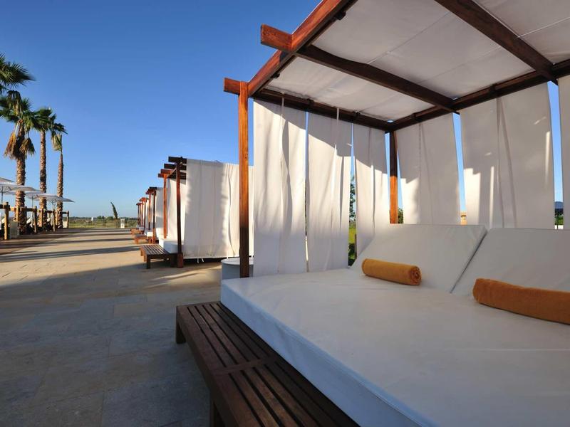 Strandliegen mit Sonnenschutz und Kissen, blauer Himmel, Palmen, ruhiger Sandweg.