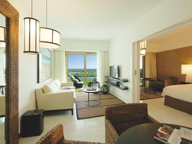 Helles, modernes Wohnzimmer mit Sofa, Sesseln, großem Spiegel und angrenzendem Schlafzimmer.