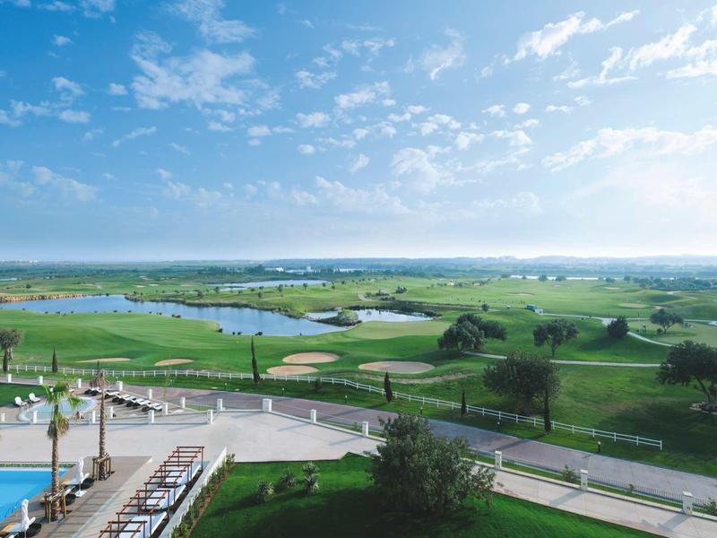 Weite grüne Golfplatzlandschaft mit kleinen Teichen, Bäumen und blauem Himmel mit Wolken.