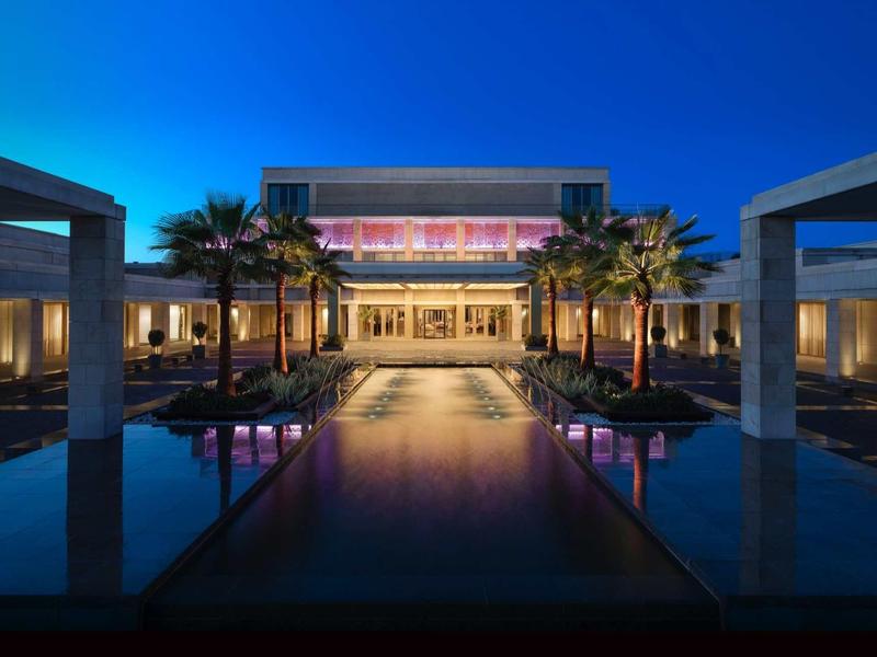 Moderner Eingangsbereich mit beleuchteter Wasserfläche, Palmen und klarer Architektur bei Abenddämmerung.