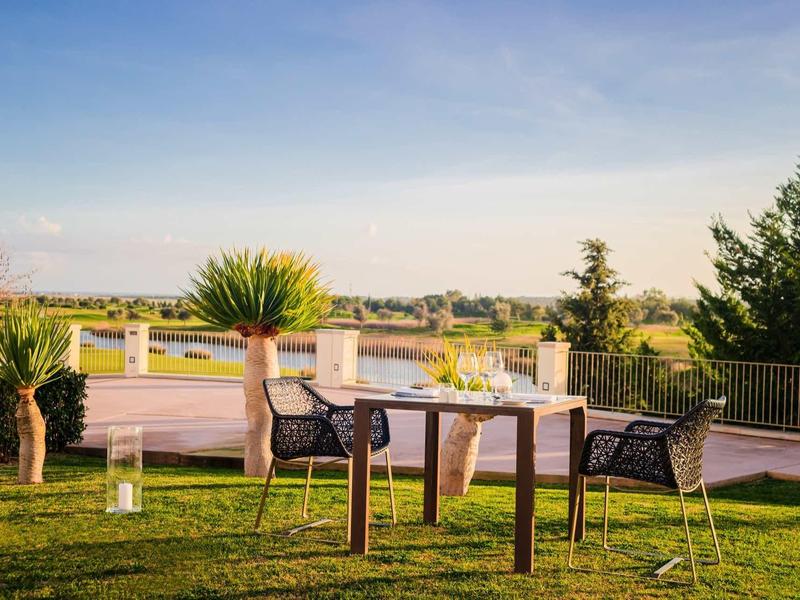 Tisch mit zwei Stühlen auf grüner Wiese, umgeben von Pflanzen und weitem Ausblick unter blauem Himmel.