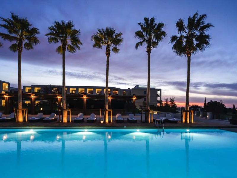 Großer beleuchteter Pool vor einem Hotel mit Palmen bei Dämmerung und blauem Himmel.