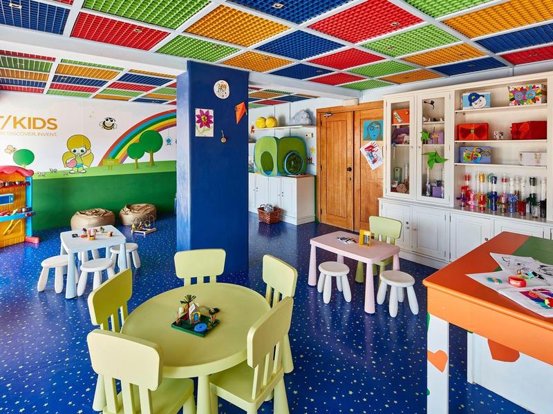 Bunter Kindergartenraum mit kleinen Tischen, Stühlen, Regalen und kindgerechter Dekoration.