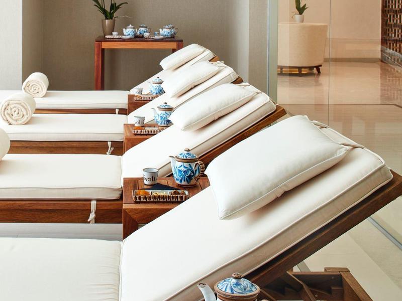 Weiße Liegen mit Kissen und blauen Teekannen stehen in einem hellen Wellnessraum aus Holz und Glas.