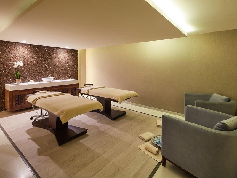 Helles Wellnesszimmer mit zwei Massagebetten, zwei grauen Sesseln und gemütlicher Beleuchtung.