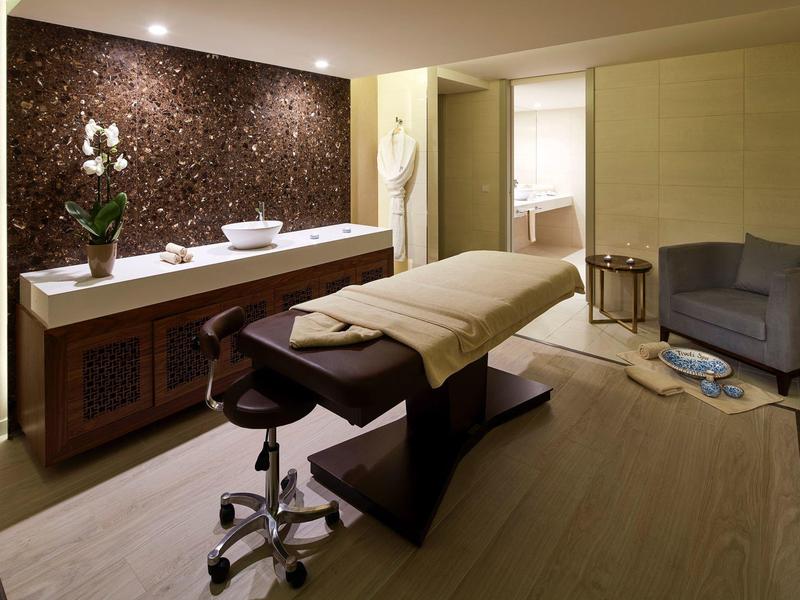 Modern eingerichteter Wellnessraum mit Massageliege, Pflanzen, grauem Sessel und gedämpfter Beleuchtung.