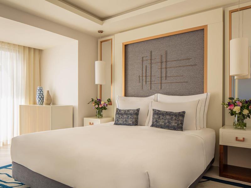 Helles Schlafzimmer mit großem Bett, gepolstertem Kopfteil, weißen Vorhängen und Blumen auf Nachttischen.