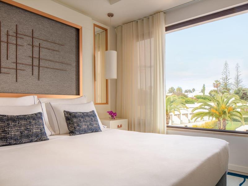 Helles Schlafzimmer mit großem Bett, moderner Wanddeko und großem Fenster mit Blick auf Palmen.