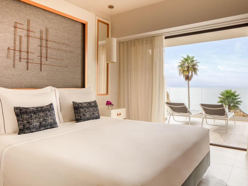 Modernes Schlafzimmer mit großem Bett, Kunst über Kopfteil und Terrasse mit Meerblick und Palmen.