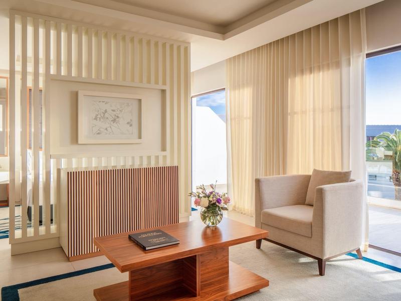 Helles Hotelzimmer mit modernem Mobiliar, Sitzbereich und Blick auf Terrasse mit Palmen.