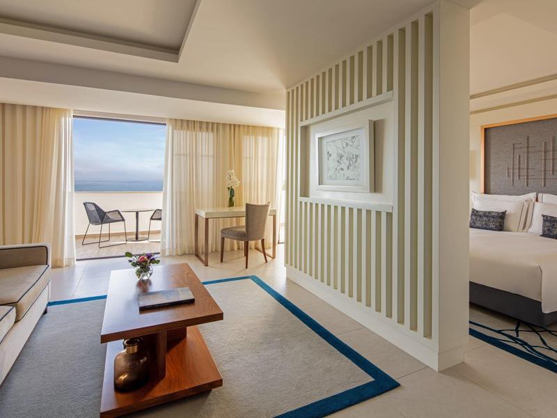 Helles Hotelzimmer mit Wohnbereich, Schlafzimmer, Teppichboden und Balkon mit Meerblick.