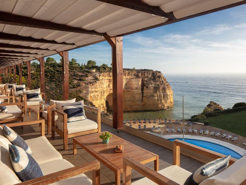 Terrassenlounge mit Holzmöbeln, Meerblick, Pool und Felsklippen im Sonnenlicht.