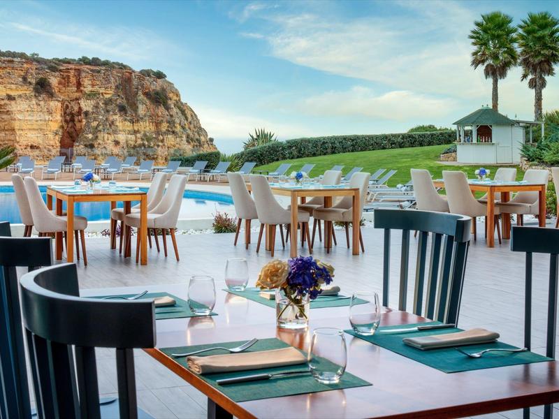 Terrasse mit Tischen und Stühlen, Blick auf Pool, Palmen und Felsen unter blauem Himmel.