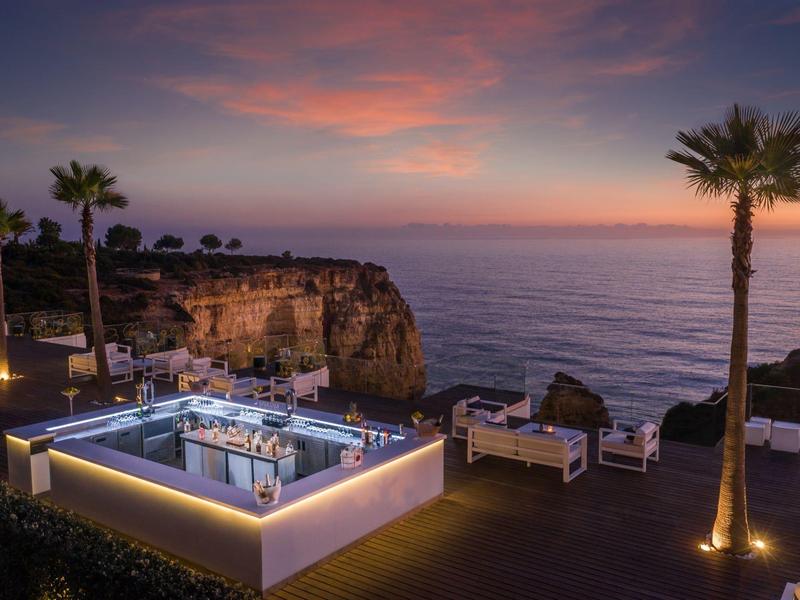 Luxuriöse Terrasse mit Pool und Palmen bei Sonnenuntergang am Meer mit Felsen in der Ferne.