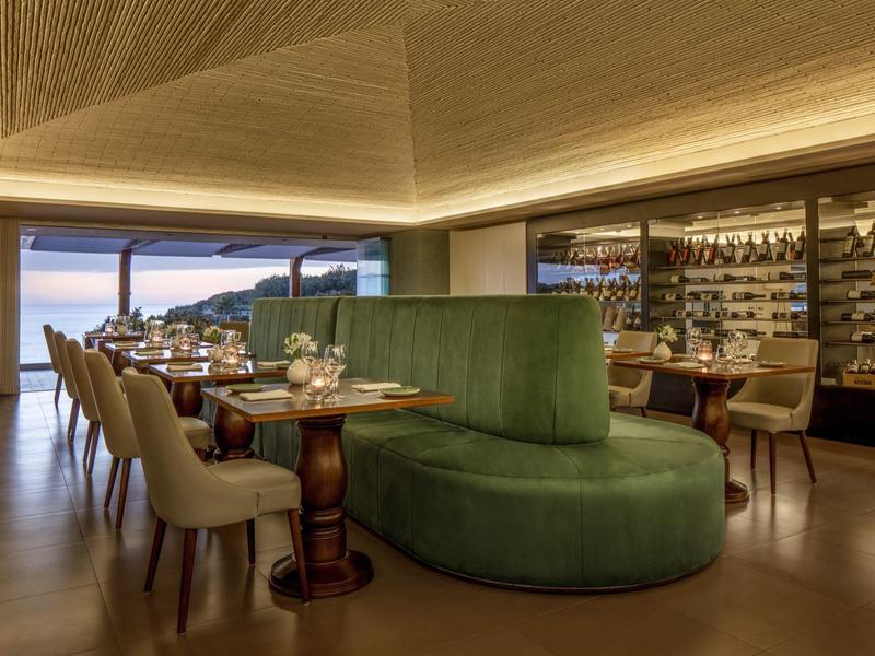 Moderner Restaurantbereich mit grünem Samtsofa, Holztischen, beigen Stühlen und Weinschrank im Hintergrund.