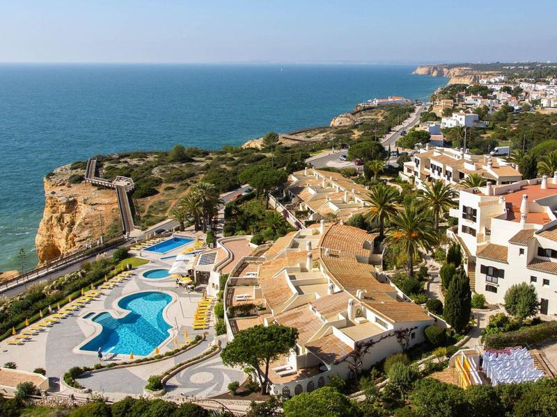Küstenort mit Poolanlage, umliegenden Häusern und grünen Felsen am blauen Meer.