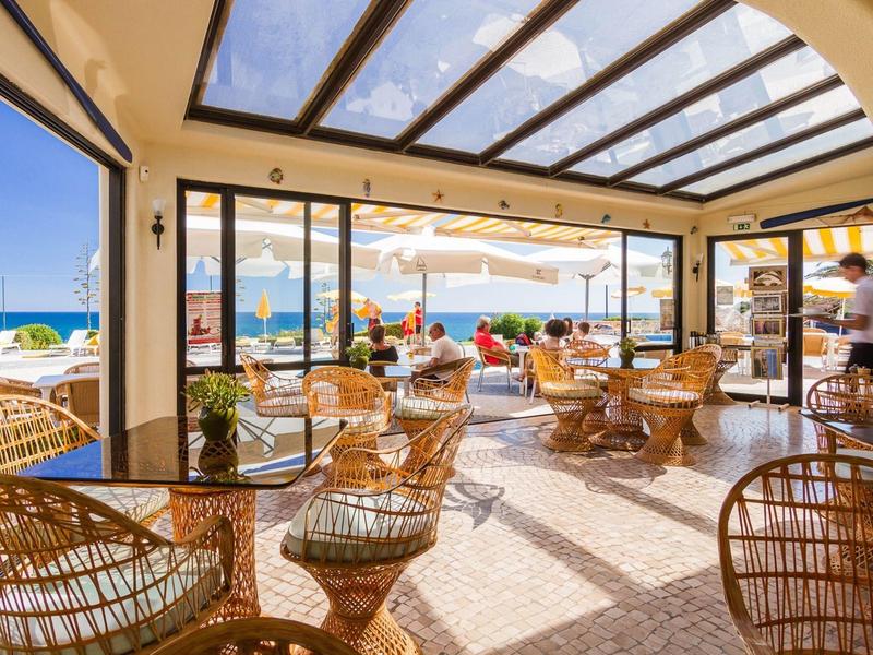 Helles Café mit Rattanstühlen, Glasdach und Blick auf Meer und Strand bei Sonnenschein.