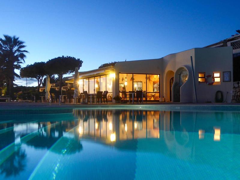 Abendliche Villa mit beleuchtetem Innenraum, Pool im Vordergrund und Palmen am Rand.