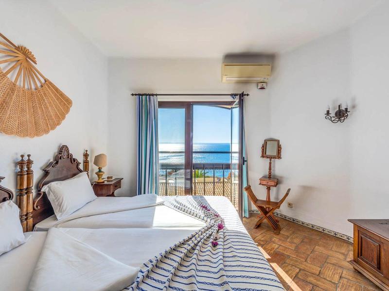 Helle Schlafzimmer mit zwei Betten, Holzmöbeln, blauen Vorhängen und Blick auf das Meer.