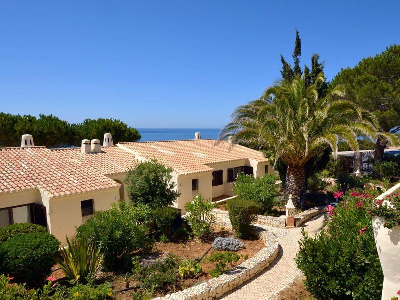 Haus mit rotem Ziegeldach, umgeben von grünen Pflanzen und Palmen unter klarem, blauem Himmel.
