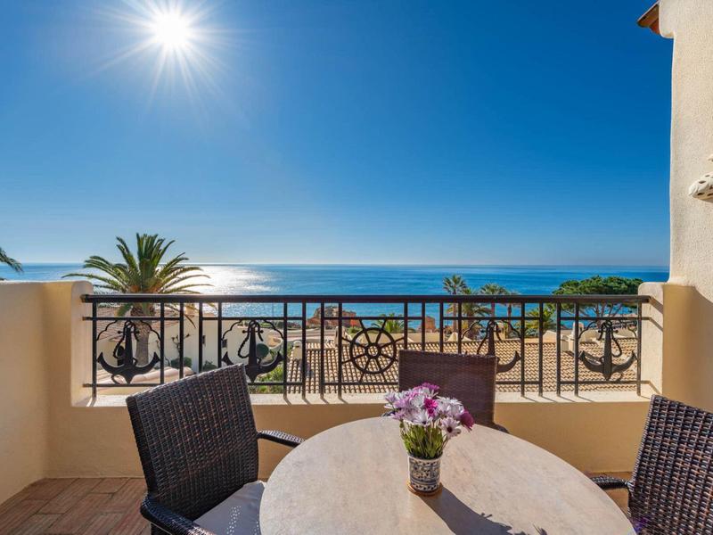 Balkon mit Tisch, Stühlen und Blumen, Blick auf Meer unter klarem blauem Himmel und hellem Sonnenlicht.