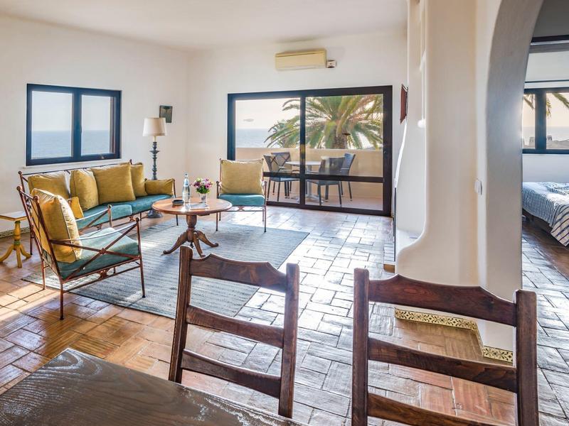 Helles Wohnzimmer mit gelbem Sofa, Glastisch, Terrakottaboden und Blick auf Terrasse mit Palme.