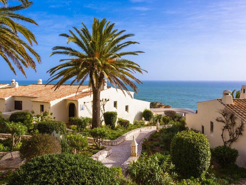 Strandnahe Häuser mit roten Dächern, Palmen und üppigem Grün bei blauem Himmel und Meer im Hintergrund.