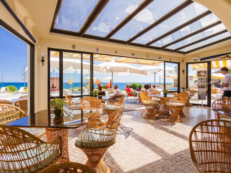 Heller Frühstücksraum mit Korbmöbeln, Glasdach und Blick auf Terrasse und Meer am sonnigen Tag.