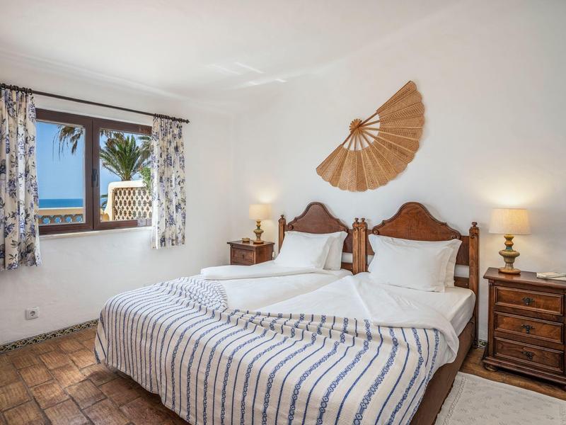 Helles Schlafzimmer mit Holzbetten, blauen Vorhängen, Ventilator-Deko und Meeresblick durchs Fenster.