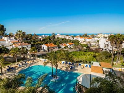Großer blauer Pool mit Palmen, grünen Rasen, weißen Häusern und blauem Himmel am Meer im Hintergrund.