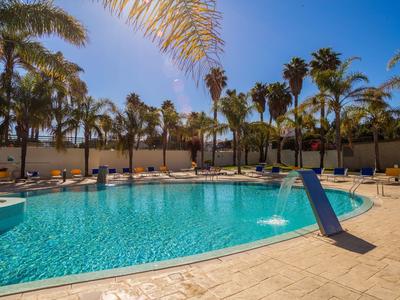 Großer, blauer Pool mit Palmen rundherum und Sonnenliegen auf sandfarbenem Boden.