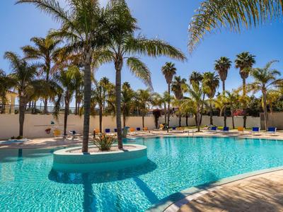 Großer Pool mit klarem blauem Wasser, umgeben von Palmen und Liegestühlen unter blauem Himmel.