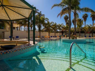 Einladender Pool mit klar blauem Wasser, umgeben von Palmen und Sonnenschirmen unter blauem Himmel.