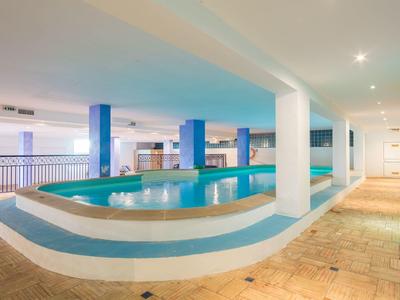 Innenraum mit rundem Schwimmbecken, blauen Säulen und beigefarbenem Fliesenboden, helle Beleuchtung.