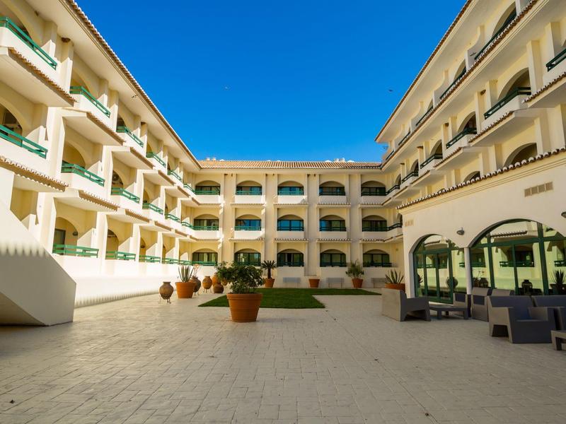 Innenhof eines mehrstöckigen Hotels mit weißen Wänden, blauen Sonnenschirmen und blauen Himmel.