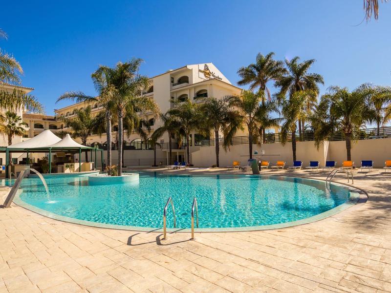 Großer Pool mit klar blauem Wasser, umgeben von Palmen und Liegestühlen unter klarem Himmel.