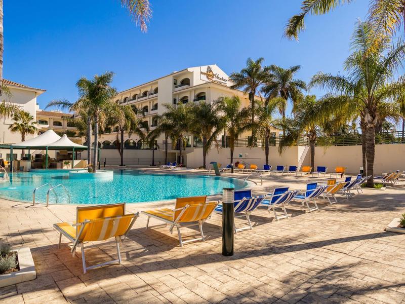 Hotelpool mit Sonnenliegen und Palmen unter klarem blauem Himmel an sonnigem Tag.